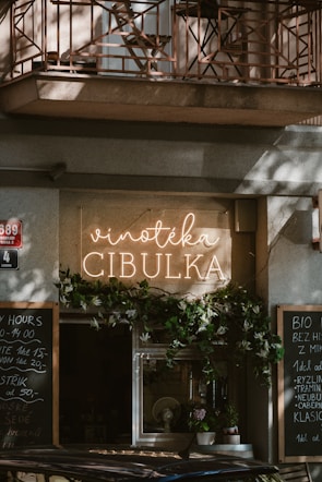 A cozy storefront features a sign with 'Vinotéka Cibulka' in glowing letters. The entrance is adorned with greenery, creating an inviting atmosphere. Chalkboards with writing are positioned on either side, listing various items or daily specials. The architecture includes a balcony with a decorative iron railing above.