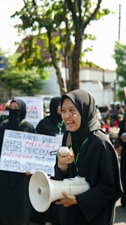 A group of people are gathered in an outdoor setting, with one person in the foreground smiling broadly. They are holding a megaphone, and have face paint of a flag on their cheek. Other people in the background are holding signs with text. Trees and buildings are visible in the background, suggesting a public or urban environment.