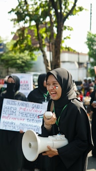 A group of people are gathered in an outdoor setting, with one person in the foreground smiling broadly. They are holding a megaphone, and have face paint of a flag on their cheek. Other people in the background are holding signs with text. Trees and buildings are visible in the background, suggesting a public or urban environment.