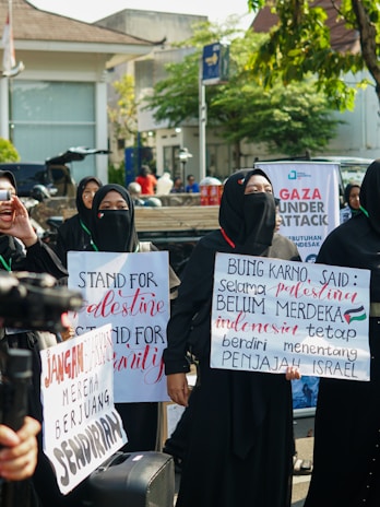 A group of individuals are standing outdoors, wearing black clothing and hijabs. They are holding signs with messages of support for Palestine. The background shows a street scene with trees and buildings.