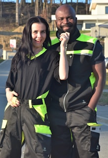 A man and a woman are smiling and standing together wearing matching dark work uniforms with bright green accents. They are outdoors on a sunny day, in what appears to be an industrial or residential area.