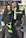 A man and a woman are smiling and standing together wearing matching dark work uniforms with bright green accents. They are outdoors on a sunny day, in what appears to be an industrial or residential area.
