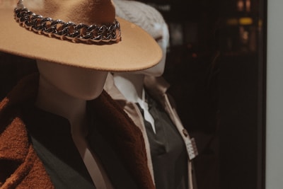 Two mannequins are showcased wearing elegant clothing. The foreground mannequin is adorned with a beige hat featuring a metallic chain detail and a brown coat over a dark top. The mannequin in the background is dressed more casually, with a light-colored coat and a knit hat.