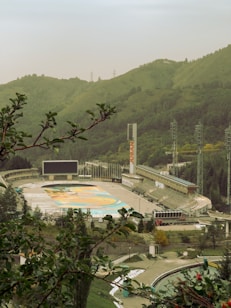 An outdoor stadium surrounded by lush green hills and forests. The seating area and playing field are visible, with a large screen and lighting towers on the sides. The scene is framed by tree branches in the foreground, enhancing the natural setting.