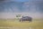 A large hippopotamus walks across a grassy plain with a safari vehicle in the background. The scenery features open grasslands with a distant view of rugged hills under a clear blue sky.
