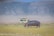 A large hippopotamus walks across a grassy plain with a safari vehicle in the background. The scenery features open grasslands with a distant view of rugged hills under a clear blue sky.