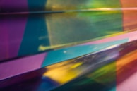 A vivid interplay of colors and light creates a layered, abstract pattern. The image features bands of blue, green, yellow, purple, and pink, blended together in a gradient. The hues shift across the composition, giving a sense of depth and movement.