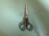A pair of scissors with gray handles and blue accents lies on a light blue surface. The surface shows signs of wear with various faint markings and small scratches visible around the placement of the scissors.