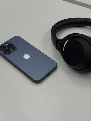 A smartphone with a greyish-blue back, displaying an Apple logo, lies on a flat, light-colored surface next to a pair of black over-ear headphones. The phone has multiple camera lenses, and the headphones feature the brand name in small text.