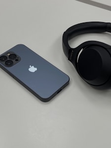 A smartphone with a greyish-blue back, displaying an Apple logo, lies on a flat, light-colored surface next to a pair of black over-ear headphones. The phone has multiple camera lenses, and the headphones feature the brand name in small text.