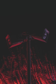 A set of loudspeakers mounted on a tall pole against a dark background. The scene is illuminated with a dramatic red light, casting shadows and creating a textured effect on the ground beneath the speakers.