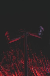 A set of loudspeakers mounted on a tall pole against a dark background. The scene is illuminated with a dramatic red light, casting shadows and creating a textured effect on the ground beneath the speakers.