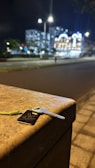 A key with a keychain lies on the edge of a stone surface by the roadside. In the background, blurred city lights and buildings can be seen, creating a bokeh effect. The scene takes place at night, with streetlights illuminating the area.