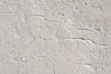 A close-up view of a textured concrete surface with visible cracks running through it. The surface appears rough and uneven, with natural imperfections and small specks of color scattered throughout.