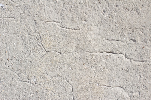 A close-up view of a textured concrete surface with visible cracks running through it. The surface appears rough and uneven, with natural imperfections and small specks of color scattered throughout.