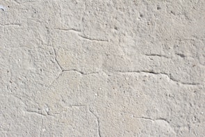 A close-up view of a textured concrete surface with visible cracks running through it. The surface appears rough and uneven, with natural imperfections and small specks of color scattered throughout.
