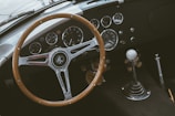 A classic car interior featuring a wooden steering wheel with the AC logo at its center. The dashboard displays a collection of vintage dials and gauges, including a speedometer and tachometer. A gear shift with a white knob is positioned on the right.