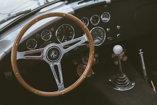 A classic car interior featuring a wooden steering wheel with the AC logo at its center. The dashboard displays a collection of vintage dials and gauges, including a speedometer and tachometer. A gear shift with a white knob is positioned on the right.