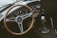A classic car interior featuring a wooden steering wheel with the AC logo at its center. The dashboard displays a collection of vintage dials and gauges, including a speedometer and tachometer. A gear shift with a white knob is positioned on the right.