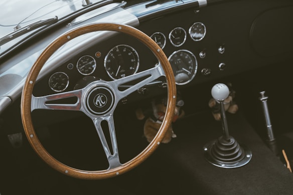 A classic car interior featuring a wooden steering wheel with the AC logo at its center. The dashboard displays a collection of vintage dials and gauges, including a speedometer and tachometer. A gear shift with a white knob is positioned on the right.