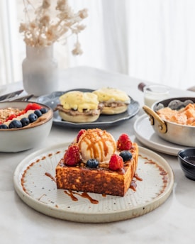 A close-up of a stylish breakfast or brunch setting featuring a thick slice of toast topped with a scoop of vanilla ice cream, drizzled with caramel sauce, and adorned with fresh raspberries and blueberries. Surrounding dishes include a bowl of acai with granola, sliced fruit, and garnishes, a plate of eggs benedict, and a dish with sweet potatoes or a similar side. The background is bright with natural lighting coming through a sheer curtain, and a decorative vase with dried flowers adds a touch of elegance.