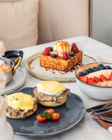 A table is set with an assortment of brunch dishes including a thick slice of French toast topped with vanilla ice cream, berries, and drizzled with caramel sauce. Nearby, a bowl contains yogurt with layers of sliced bananas, strawberries, blueberries, and granola. There are also two eggs benedict on a dark plate, garnished with cherry tomatoes and parsley.