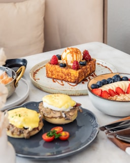 A table is set with an assortment of brunch dishes including a thick slice of French toast topped with vanilla ice cream, berries, and drizzled with caramel sauce. Nearby, a bowl contains yogurt with layers of sliced bananas, strawberries, blueberries, and granola. There are also two eggs benedict on a dark plate, garnished with cherry tomatoes and parsley.
