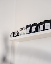 A minimalistic shelf displays several black packages of Dot Cafe coffee, each labeled with different origins such as Colombia and India. An old-fashioned black clock with two faces sits alongside, adding a touch of retro charm. The word 'COFFEE' is partially visible in black letters on the white wall.
