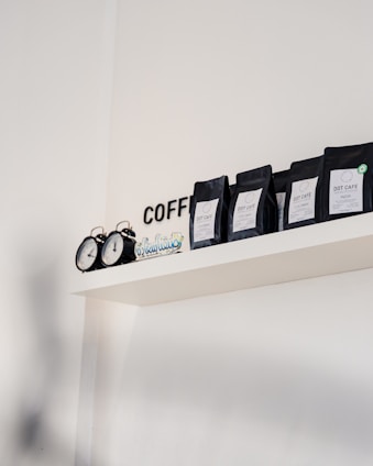 A minimalistic shelf displays several black packages of Dot Cafe coffee, each labeled with different origins such as Colombia and India. An old-fashioned black clock with two faces sits alongside, adding a touch of retro charm. The word 'COFFEE' is partially visible in black letters on the white wall.