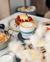 A café table setting with a focus on a cup of latte art, accompanied by various dishes including a waffle topped with ice cream, berries, and caramel sauce, beside a dish with a golden handle.
