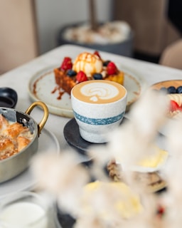 A café table setting with a focus on a cup of latte art, accompanied by various dishes including a waffle topped with ice cream, berries, and caramel sauce, beside a dish with a golden handle.