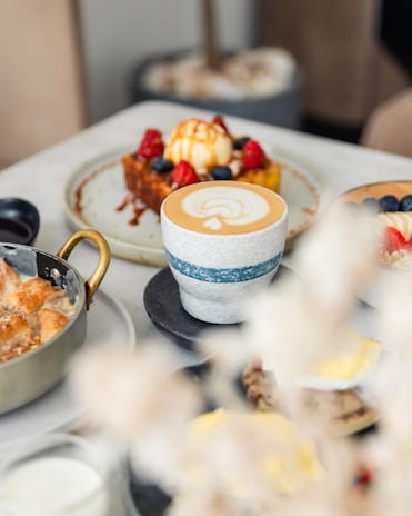 A café table setting with a focus on a cup of latte art, accompanied by various dishes including a waffle topped with ice cream, berries, and caramel sauce, beside a dish with a golden handle.