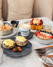 A table set with a variety of brunch items, including a ceramic cup with cappuccino and latte art, eggs Benedict on English muffins with hollandaise sauce, a bowl of granola topped with fresh strawberries, blueberries, and banana slices, and a dessert dish garnished with raspberries, blueberries, and drizzled caramel. A small skillet with pastries is also present, alongside two halves of cherry tomatoes.