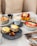 A table set with a variety of brunch items, including a ceramic cup with cappuccino and latte art, eggs Benedict on English muffins with hollandaise sauce, a bowl of granola topped with fresh strawberries, blueberries, and banana slices, and a dessert dish garnished with raspberries, blueberries, and drizzled caramel. A small skillet with pastries is also present, alongside two halves of cherry tomatoes.