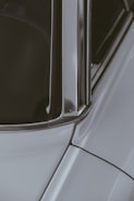 A close-up section of a car showing part of the windshield, window frame, and roof with smooth metal and glass surfaces reflecting light. The structured lines and edges of the car's bodywork are prominent.