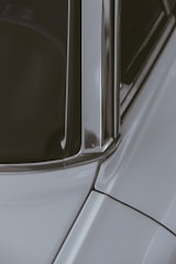 A close-up section of a car showing part of the windshield, window frame, and roof with smooth metal and glass surfaces reflecting light. The structured lines and edges of the car's bodywork are prominent.
