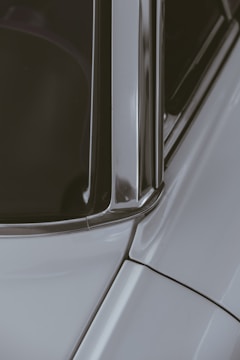 A close-up section of a car showing part of the windshield, window frame, and roof with smooth metal and glass surfaces reflecting light. The structured lines and edges of the car's bodywork are prominent.