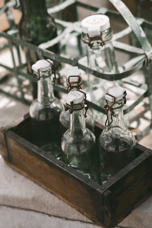 Several glass bottles with swing-top caps are neatly arranged in a wooden crate. The crate is placed on a rustic surface with a metal bottle rack visible in the background. The scene is illuminated by soft, natural lighting, creating muted reflections on the glass surfaces.