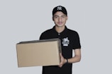 A person wearing a black cap and a black polo shirt with a courier service logo holds a large cardboard box. The background is a plain light gray, and the person appears to be smiling slightly.