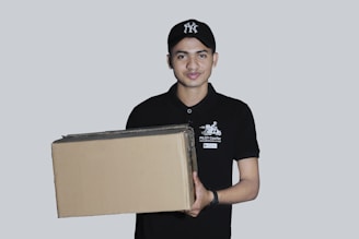 A person wearing a black cap and a black polo shirt with a courier service logo holds a large cardboard box. The background is a plain light gray, and the person appears to be smiling slightly.