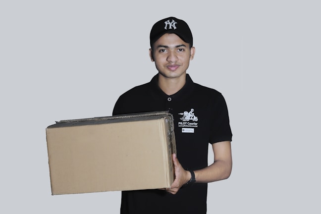 A person wearing a black cap and a black polo shirt with a courier service logo holds a large cardboard box. The background is a plain light gray, and the person appears to be smiling slightly.