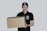 A person wearing a black cap and a black polo shirt with a courier service logo holds a large cardboard box. The background is a plain light gray, and the person appears to be smiling slightly.