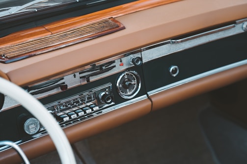 A vintage car dashboard featuring a classic radio with tuning dials and buttons. The dashboard is made of brown leather and wood, with a smooth and polished finish. The design includes chrome accents and an analog clock.