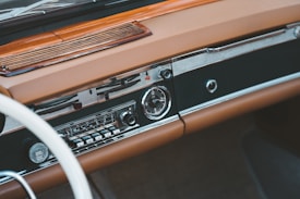 A vintage car dashboard featuring a classic radio with tuning dials and buttons. The dashboard is made of brown leather and wood, with a smooth and polished finish. The design includes chrome accents and an analog clock.