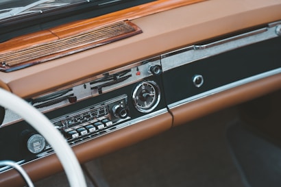 A vintage car dashboard featuring a classic radio with tuning dials and buttons. The dashboard is made of brown leather and wood, with a smooth and polished finish. The design includes chrome accents and an analog clock.
