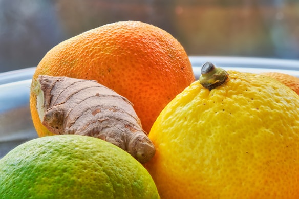 A close-up view of fresh citrus fruits including an orange, a lemon, and a lime, alongside a piece of ginger. The fruits are placed on a reflective surface with a soft-focus background.