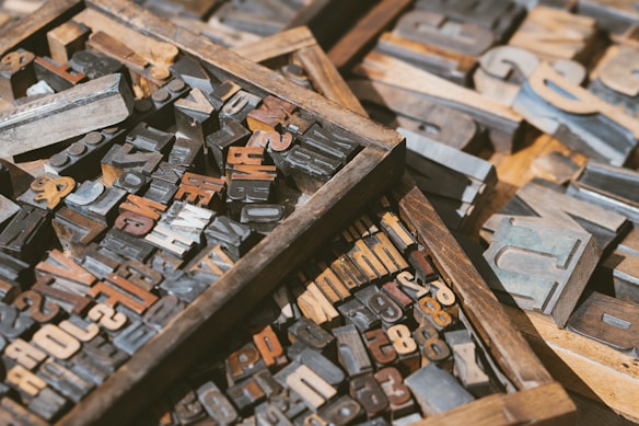 Wooden trays filled with various vintage letterpress blocks in different sizes and fonts. The blocks are arranged in a seemingly random order and feature a range of letters and numbers. The surface and edges of the blocks show signs of wear, indicating they have been used. The scene is well-lit, highlighting the textures and colors of the aged wood and metal.