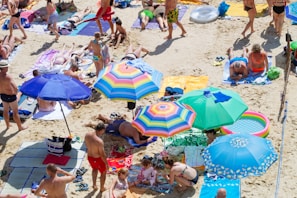 A crowded beach scene with numerous people sunbathing on colorful towels and under a variety of vibrant umbrellas. The sand is light beige, and the beachgoers, wearing swimsuits, are lounging and walking around. Inflatable pool toys and beach accessories are scattered around, including a rainbow-colored inflatable ring.