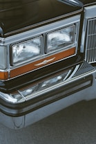 Close-up of the front end of a classic car, showcasing the headlights and a chrome grille with a vintage emblem. The headlights are rectangular with multiple beams, framed with polished chrome accents. The surface appears clean and well-maintained with a black and silver color scheme.