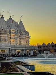 An intricately carved temple made from white stone stands against a backdrop of a warm, golden sky at sunset. The temple is adorned with decorative spires and flags. In front of the temple is a reflective water feature with a small fountain, surrounded by manicured gardens. People are gathered near the entrance, suggesting a place of worship or cultural significance.